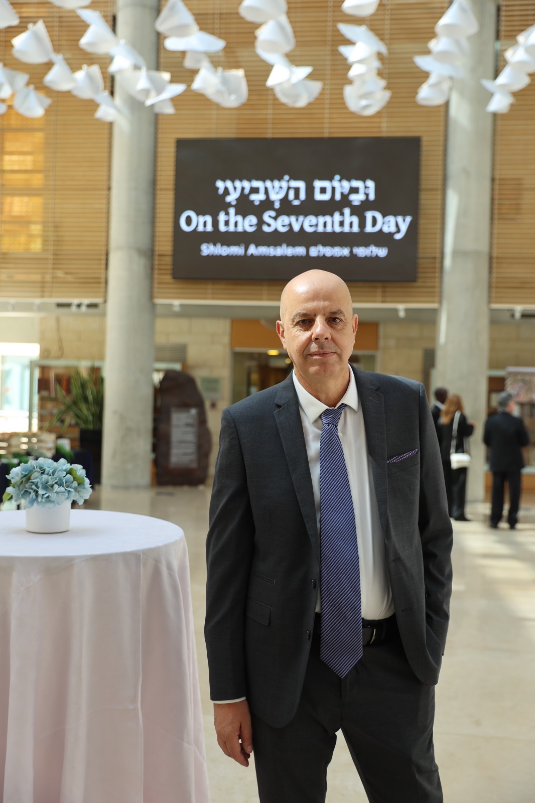 משתתף בטקס ההשקה במשרד החוץ, על רקע שלט הספר ״וביום השביעי״
